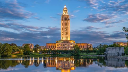 A tall, illuminated building stands by a lake at dusk, reflecting in the water. Green trees and a partly cloudy sky surround the scene, creating a peaceful cityscape.